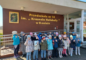 Dzieci stoją pod budynkiem przedszkola, machają rączkami, jedno z dzieci trzyma w ręku maskotkę Krasnala Hałabały.